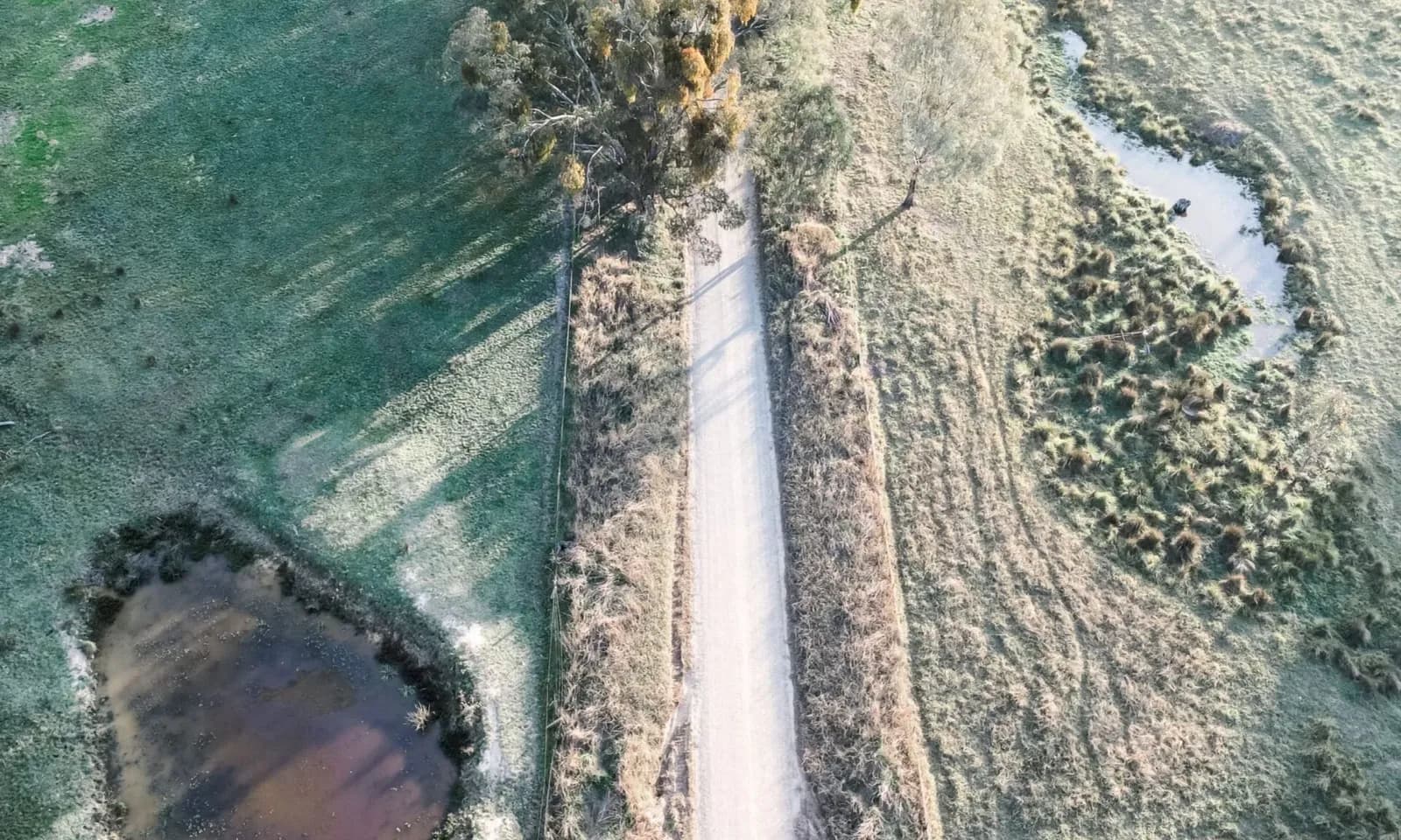 Aerial view of a dirt road running through grassy fields with trees and a pond, photographed by Hamlet & Fields