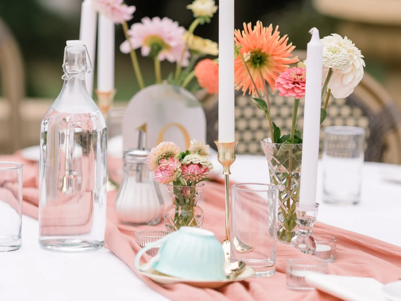 A styled table with white candles and dahlias in glass vases on a blush linen runner