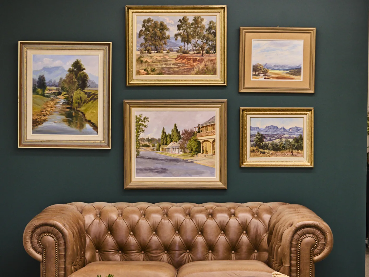 Gold-framed landscape paintings on a deep green wall above a tan leather sofa